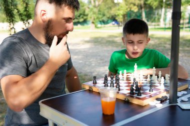 Bir adam ve bir çocuk açık havada satranç oynuyorlar. Aydınlık ve güneşli bir günde ortam bir parktır. Odak noktası onların etkileşimi ve satranç tahtası..