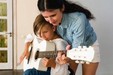 İlgili bir yetişkin gitar öğrenen genç bir çocuğa yardım eder. Eğitmenin ellerini gösteren yakın çekim beyaz akustik gitarla öğrenciye nazikçe rehberlik ediyor..