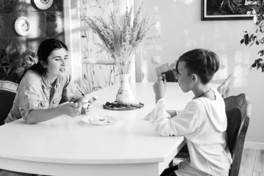 Gri tonlu bir görüntü, güneşli bir odada beyaz bir masada oturan iki çocuğu yakalıyor. Kardeş gibi görünüyorlar, sessiz bir etkileşimi paylaşıyorlar..