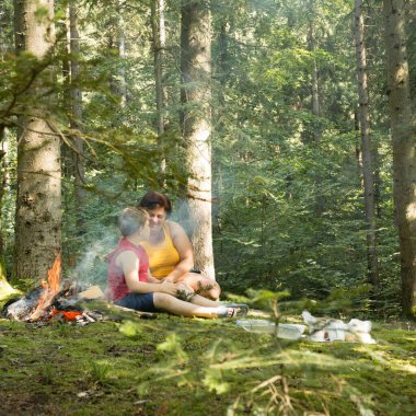 Bir kadın ve küçük bir çocuk, yemyeşil bir ormanda, küçük bir kamp ateşinin yanında, ağaçların arasında birlikte sakin bir anı paylaşıyorlar. Güneş ışığı yaprakların arasından süzülür..