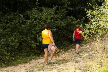 Bir kadın ve bir çocuk ormanda bir patikada yürüyüş yapıyorlar. Kadın bir şapka taşıyor ve kameradan uzaklaşıyorlar..