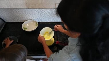 Parlak bir mutfakta, bir anne, çocuğu hevesle izlerken maharetli bir şekilde yumurta pişirir ve paylaşılan yemek anlarından oluşan neşeli bir atmosfer yaratır..