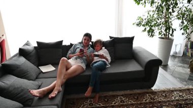 Bir anne ve küçük oğlu rahat gri bir kanepede rahat bir şekilde dinlenir. Parlak, bitki dolu oturma odalarında ekran başında eğlenirken gülümserler..