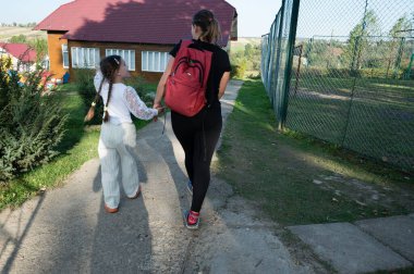 Kırmızı sırt çantalı bir anne, bazı binalara doğru giden bir yolda kızıyla el ele tutuşup uzaklaşır. Geçidin yanında zincir bir çit var, uzaktaki ağaçlar..