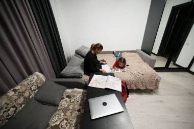 Bu samimi görüntü, bir anne ve kızın evde kaliteli zaman geçirdiklerini gösteriyor. Anne, kız çizim yaparken bir kitap okuyor..