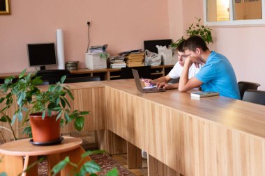 İki öğrenci bir sınıfta büyük, hafif bir ahşap masada dizüstü bilgisayarda birlikte çalışıyorlar. Yakınlarda bir saksı bitkisi oturuyor. Kitaplar ve bilgisayar ekipmanları arka planda, oda ışıkla dolu..