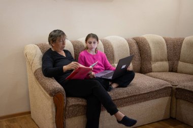 Bir kadın bir kitapla otururken, bir kız sıcak bir evdeki kanepede dizüstü bilgisayar kullanıyor. Nesiller boyunca iletişimi ve işbirliğini teşvik eden bir öğrenim ortamındalar..