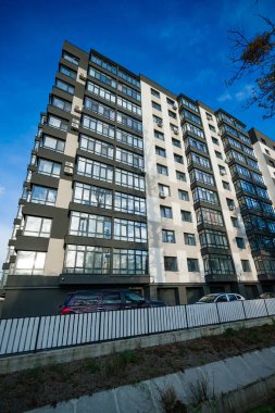 Kalın çizgiler ve geniş pencerelerle çağdaş bir apartman kompleksi yükseliyor. Güneş ışığı cam cephelerinden yansıyarak modern mimari ve şehir yaşamını ön plana çıkarır..