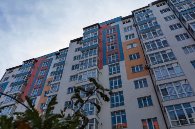 Dayanıklı yapı, gökyüzünü yansıtan geniş pencereli renkli bir cephe, yeşillik ve kentsel hayatın bir ipucunu barındıran, dimdik ayakta duruyor..