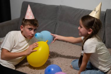 Parti şapkalı iki çocuk, rahat bir oturma odasında yumuşak bir halıda renkli balonlarla oynuyorlar. Neşeli bir anda gülümseme ve kahkahalar atıyorlar..