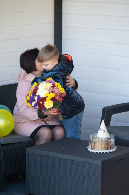 Bir anne ve küçük oğlu birlikte kutlarken kalpten gelen bir an. Çocuk bir çiçek buketi sunar, etrafında balonlar ve bir pasta vardır..