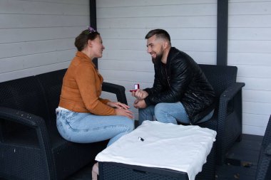 Sakin bir açık hava köşesinde, bir adam eşine bir yüzük sunar, neşe, sevgi ve içten bir beklentiyle dolu samimi bir an yaratır..