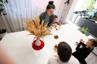 Bir aile sıcak içecekler ve hamur işlerinin keyfini çıkarırken, parlak mutfak ortamları paylaşılan bir yemek ve güzel bir birliktelik üzerine anılar yaratıyor..