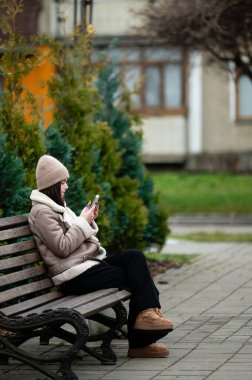 Genç bir kadın rahat bir şekilde ahşap bir bankta oturuyor, telefonuna dalmış, şehirdeki sakin bir akşam gökyüzünün altında yemyeşil yeşermiş, sükunet ve rahatlığı yansıtıyor..