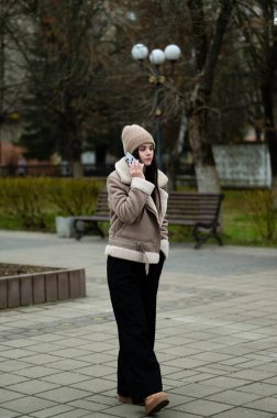 Serin bir sonbahar gününde genç bir kadın parkta telefon görüşmesi yapıyor. Etrafı ağaçlar ve banklarla çevrili rahat bir palto ve şapka giyiyor..