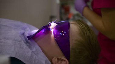 Bir diş hekimi klinik ortamda ağır çekimde oral tedavi uygularken küçük bir çocuk, koruyucu gözlük takarak dişçi koltuğunda dinlenir..