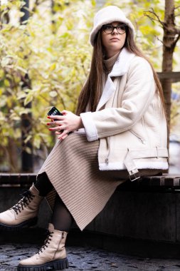 Açık havada sakin bir anın tadını çıkaran şık bir birey rahat bir bankta oturur, sıcak bir takım elbise, gözlük ve canlı bir düşüşün ortasında bir şapka giyer..