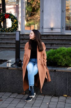 Şık bir palto giymiş genç bir kadın, bir bankta rahat bir şekilde oturur, güzel bir tatil süsü alır ve kış soğuğunun ortasında onu çevreleyen parlak ışıkları yakar..