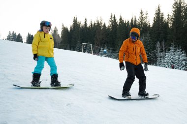 İki çocuk parlak kış kıyafetleri içinde karlı bir yamaçta ustalıkla geziniyor. Arkaplanda sonsuz yeşil ağaçlar ve açık bir gökyüzü var. Mükemmel bir kış harikalar diyarı yaratıyor..