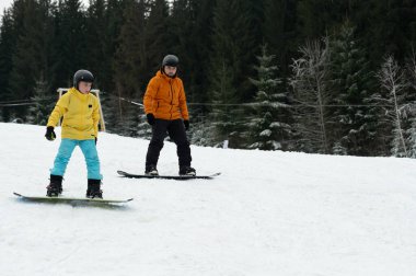 Kar sörfçüleri canlı ceketlerle karlı bir tepeden aşağı süzülüyorlar, uzun yeşil ağaçlardan oluşan bir zemin ve berrak bir kış göğüne karşı, kahkahalar ve heyecanları paylaşıyorlar..