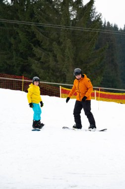 Parlak giyimli bireyler karlı bir tepede snowboard derslerinin keyfini çıkarırlar. Etrafı bulutlu bir gökyüzünün altında yeşil ağaçlarla çevriliyken gülüşmeler ve kahkahalar atarlar..
