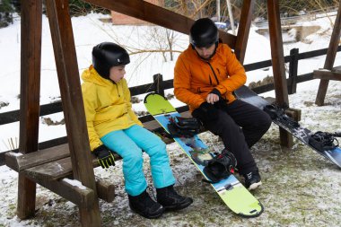 İki snowboardcu, sakin dağ manzarasıyla çevrili kar yamaçlarında neşeli bir gün için hazırlanan ahşap bir bankta oturur..