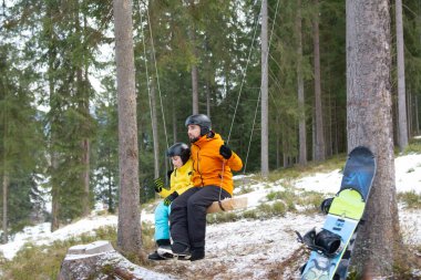 Bir adam ve bir çocuk, açık kış kıyafetleri içinde, karlı bir ormanda, ahşap bir salıncakta rahatça oturup snowboard ve eğlencenin tadını çıkarmaya hazırlar..