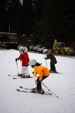 Üç çocuk karlı bir ortamda kayak aktivitelerine katılıyor. Kış günü kask ve renkli kıyafetler giyerken yeteneklerini ve heyecanlarını gösteriyorlar..