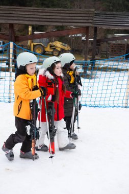 Renkli kış giysileri içinde üç hevesli çocuk, kar kaplı bir alanda, kayak ve direklerle donatılmış, kayak maceralarının tadını çıkarmaya hevesli bir şekilde bekliyorlar..