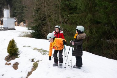 Üç çocuk miğferler ve kalın ceketler kar kaplı ormanda kış eğlencesinin tadını çıkarıyor. Soğuk bir öğleden sonra etraflarını coşkuyla ve kahkahalarla keşfediyorlar..