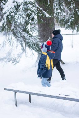 İki çocuk, kışlık giysiler içinde, karlı bir günün tadını çıkarın. Karla kaplı bir bankın yanındalar. Karla dolu bir çam ağacının altında. Sahne huzurlu ve kışlık..