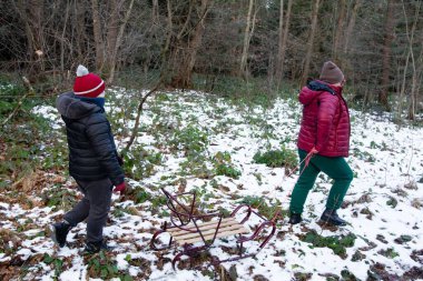 İki çocuk, karla kaplı bir orman yolunda ilerliyor. Biri eski bir ahşap kızağı çekiyor. Sahne huzurlu, doğadaki kış aktivitelerinin neşesini gösteriyor..