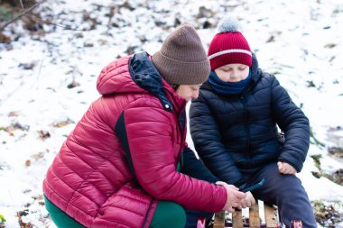 Sıcak kış giysileri giyen bir kadın ve genç bir çocuk, karlı bir çevrede ahşap bir kızağın üzerinde birlikte oturuyorlar. Akıllı bir telefona bakıyor gibi görünüyorlar..