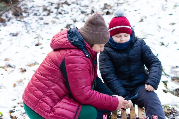 Sıcak kış giysileri giyen bir kadın ve genç bir çocuk, karlı bir çevrede ahşap bir kızağın üzerinde birlikte oturuyorlar. Akıllı bir telefona bakıyor gibi görünüyorlar..