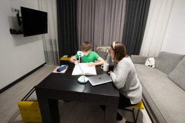 Bir anne ve oğlu ev ödevlerini şık bir oturma odasında birlikte yaparlar, akşam ışığının yumuşak sıcaklığında görevler üzerine bilgi ve yakınlaşma anlarını paylaşırlar..