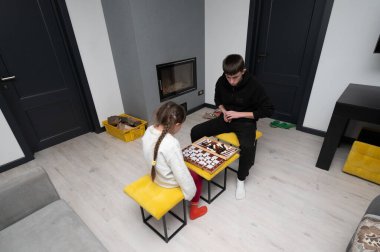 İki kardeş, sıcak oturma odalarındaki masa oyununa dikkatle odaklanmış, parlak sarı taburelerde otururken, bir şömine, sıcak bir kış öğleden sonrasının atmosferine eklenir..