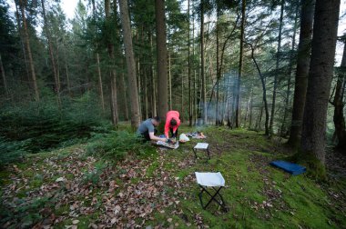 İki arkadaş, bereketli yeşilliklerin ve ormanın yükselen ağaçlarının arasında, doğanın sakinleştirici sesleriyle çevrili, açık bir ateşin üzerinde nefis bir yemek hazırlıyorlar..