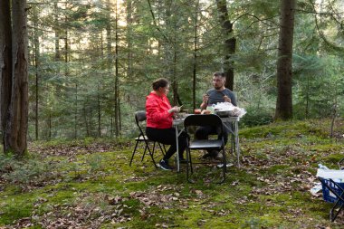 Yumuşak güneş ışığı ağaçların arasından süzülüp barışçıl bir atmosfer yaratırken iki birey yemyeşil yemliklerle çevrili küçük bir masada keyifli bir yemek yiyorlar..