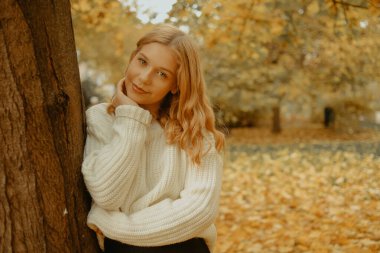 Sonbahar parkındaki kadın. Sonbahar yapraklarının arka planında genç bir kız. Beyaz, kalın kazaklı bir mankenin moda fotoğrafı. Sarışın kameraya bakıyor.