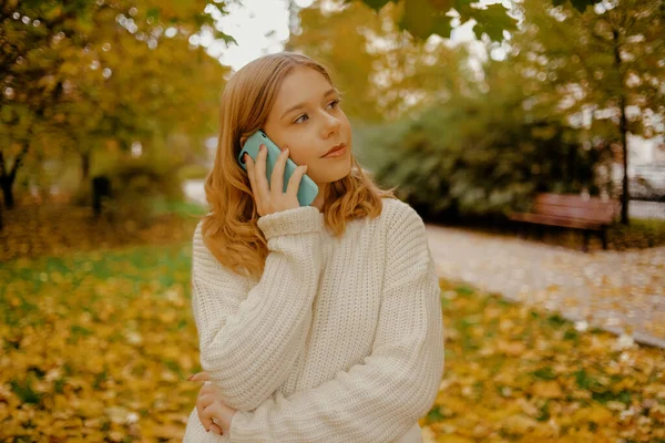 Girl teenager in the autumn park speaks by phone. A beautiful girl calls on her mobile and speaks against the background of trees.