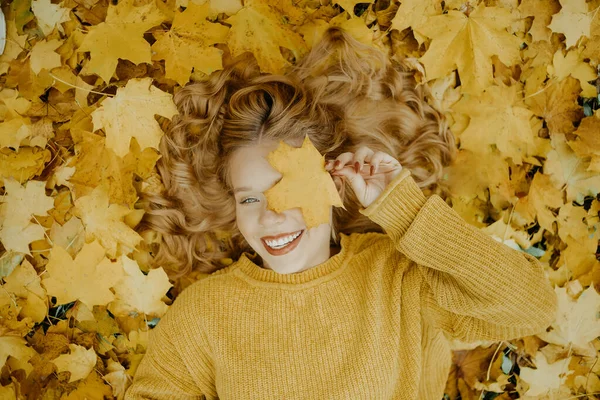 美しい少女は秋に公園にいます オレンジの葉を背景に黄色いセーターを着た10代 ブロンドは笑顔で笑う ファッションとスタイル 葉と笑顔で目を覆う女 — ストック写真