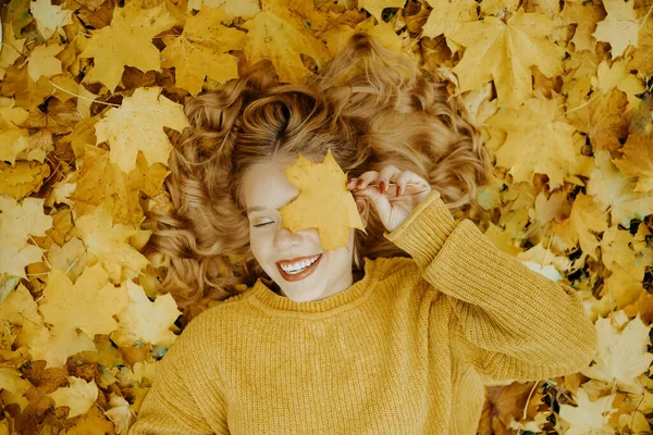 美しい少女は秋に公園にいます オレンジの葉を背景に黄色いセーターを着た10代 ブロンドは笑顔で笑う ファッションとスタイル 葉と笑顔で目を覆う女 — ストック写真