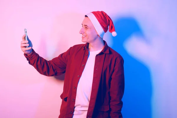 A man in a santa hat in neon light. The guy is talking by video with his family. New Year\'s and Christmas. Man with a phone in his hands