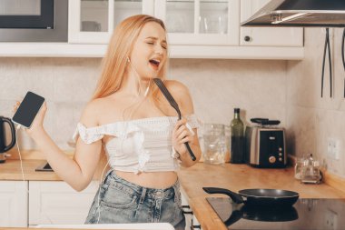  Kadın mutfakta akıllı telefon ve müzikle şarkı söylüyor ve dans ediyor. Kulaklıklı bir kız mutfakta duruyor ve indüksiyon sobasının arkasında yemek hazırlıyor..