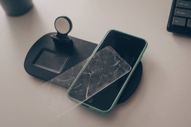 Cep telefonu masanın üzerindedir. Kırık ekranlı akıllı telefon, cam. Ekran tamiri, cam tamiri, tamir. Telefon kablosuz şarjda