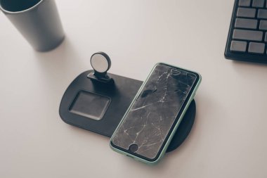 Cep telefonu masanın üzerindedir. Kırık ekranlı akıllı telefon, cam. Ekran tamiri, cam tamiri, tamir. Telefon kablosuz şarjda