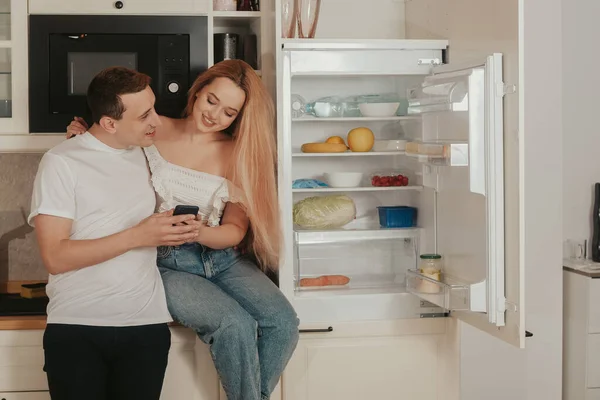 A woman and a man order food online. A couple is standing in the kitchen near the refrigerator and buying food through the phone. Online shopping, delivery.