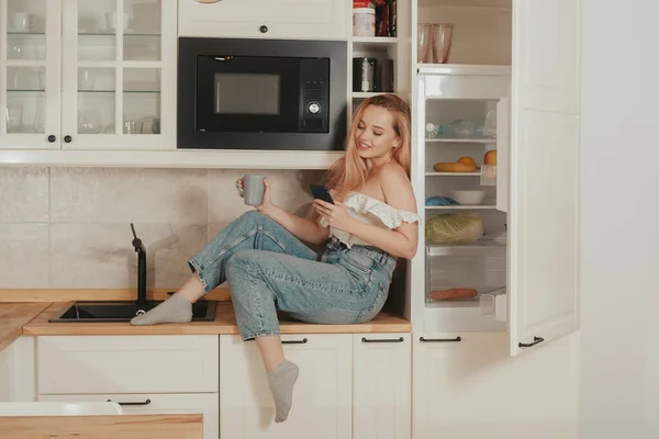 A woman is sitting at home in the kitchen at the will of the refrigerator. A girl with a good figure holds a mobile phone in her hands. Food delivery, mobile shops, online shopping. The model looks at what to order on the Internet.