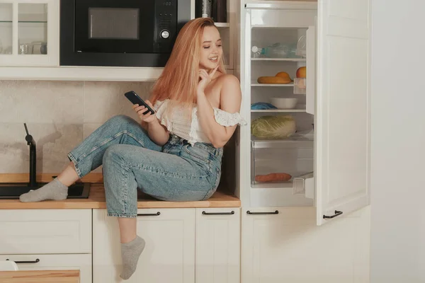 A woman is sitting at home in the kitchen at the will of the refrigerator. A girl with a good figure holds a mobile phone in her hands. Food delivery, mobile shops, online shopping. The model looks at what to order on the Internet.