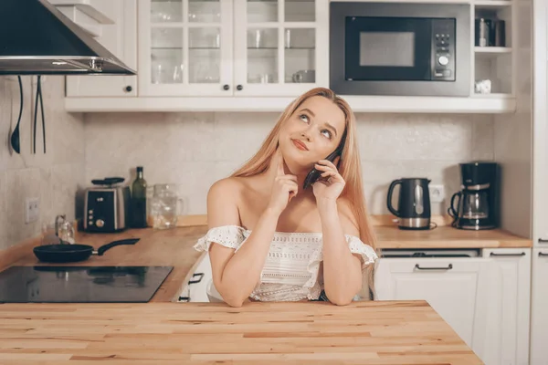Beautiful woman sits at a table on the background of the kitchen at home. The girl speaks on the phone, looks up and thinks. What to order from food in delivery.